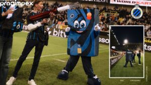 Cheerful blue square mascot posing with a large prop gun on a crowded soccer field, fans cheering in the stands, sponsor logos visible (Club Brugge crest on right).