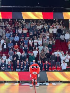 Huge crowd standing in a stadium with a red and white mascot at the front center wearing the number 13, cheering spectators behind him out of focus at the arena edge.